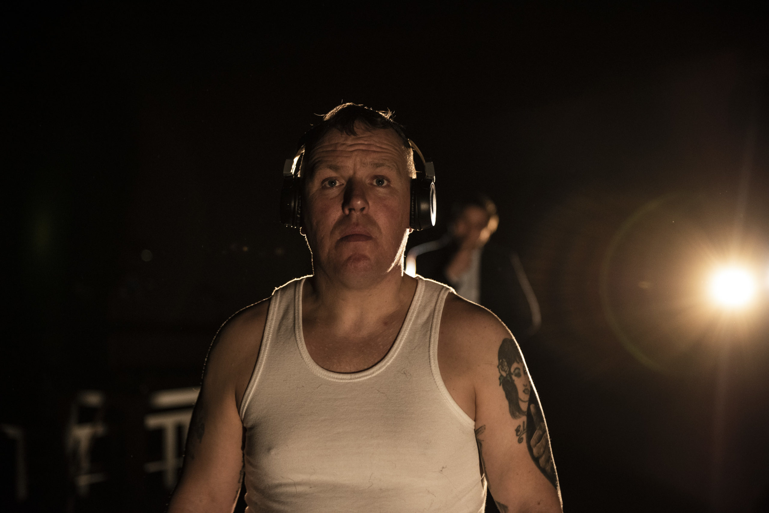 Stand up Comedian Willa White stands in a white vest and headphones on stage, behind him is Gary Keegan who speaks into a microphone in a performance of The Examination